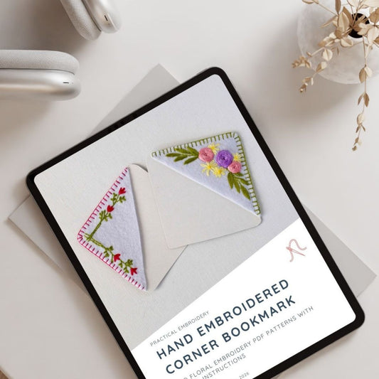 A tablet displaying an embroidery pattern cover for a hand-embroidered corner bookmark, styled on a light surface with headphones and dried flowers nearby.